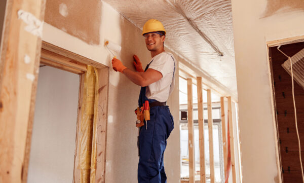 Joyful male worker in safety helmet looking at camera and smiling while painting wall with paint roller