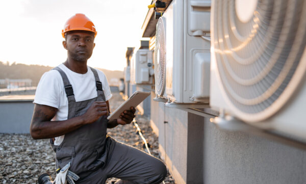 Focused man scrolling websites on digital tablet with useful information about air conditioners. African american craftsman in overalls and protective helmet looking at cooling system outdoors.